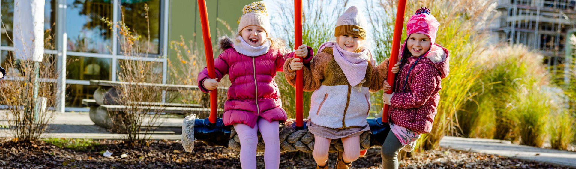 Drei Kinder sitzen auf einer Schaukel und lachen. Im Hintergrund ein Gebäude.