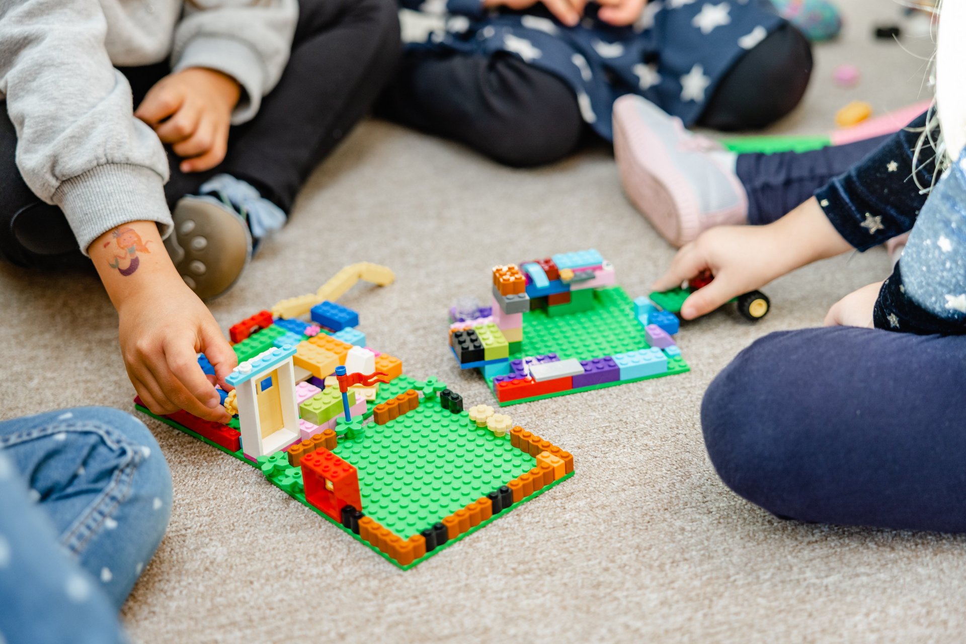 Auf dem Bild sitzen mehrere Kinder auf einem beigen Teppichboden. Sie spielen mit bunten Kunststoff-Bausteinen und bauen kleine Gebäude auf grünen Bauplatten. Zwei Bauwerke sind gut sichtbar. Die Kinderhände greifen nach Bausteinen, ein Kind hat ein temporäres Tattoo auf dem Handgelenk. Die Kleidung der Kinder ist bunt, mit Mustern wie Sternen. Die Szene zeigt konzentriertes, gemeinsames Spielen in der Kita Landwehr.